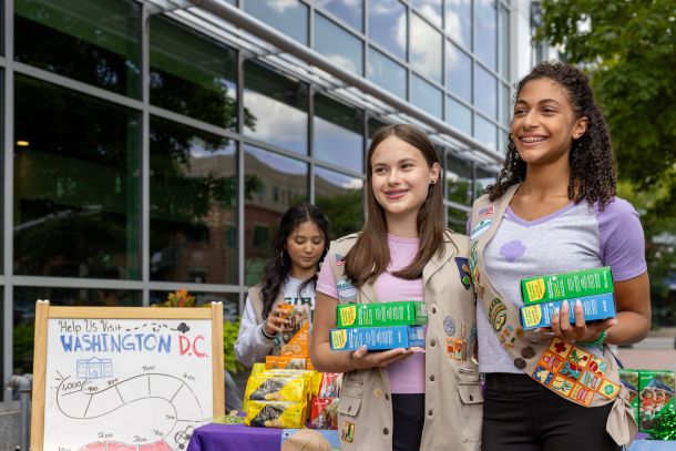 Girl Scouts holding boxes of cookies in front of a cookie booth