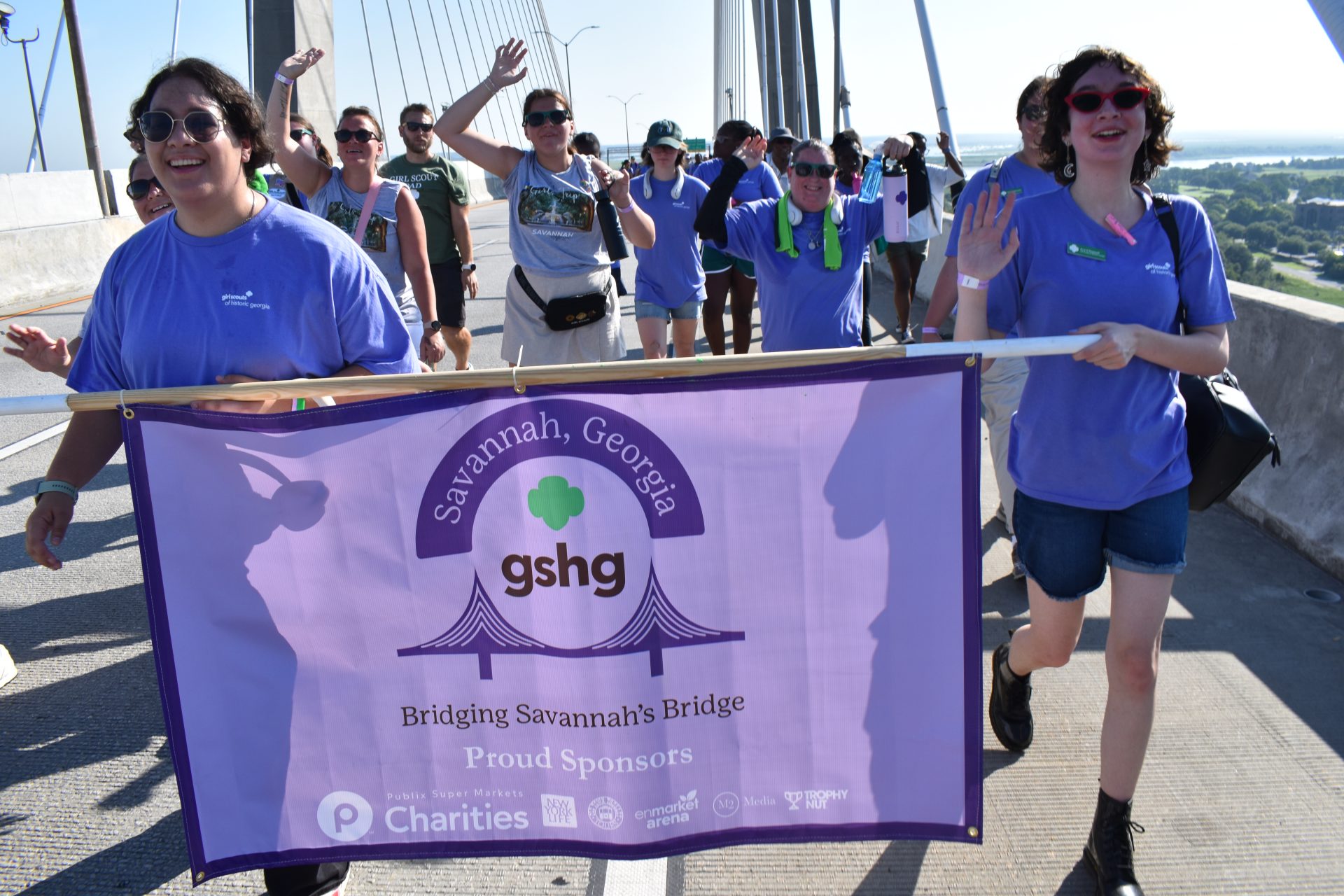 Image of Girl Scouts walking across Talmadge Bridge and holding a banner that showcases the event sponsors for Bridging Savannah's Bridge